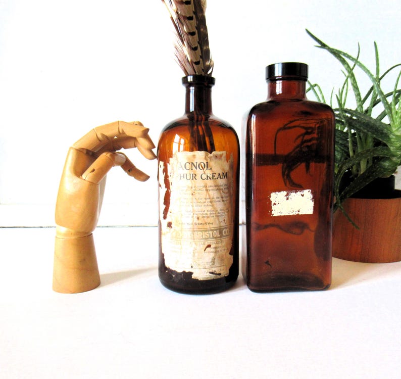 May include: Two antique amber glass bottles, one with a label reading "ACNOL HUR CREAM" and a feather, the other with a blank label. A wooden articulated hand is on the left. A potted plant is on the right.