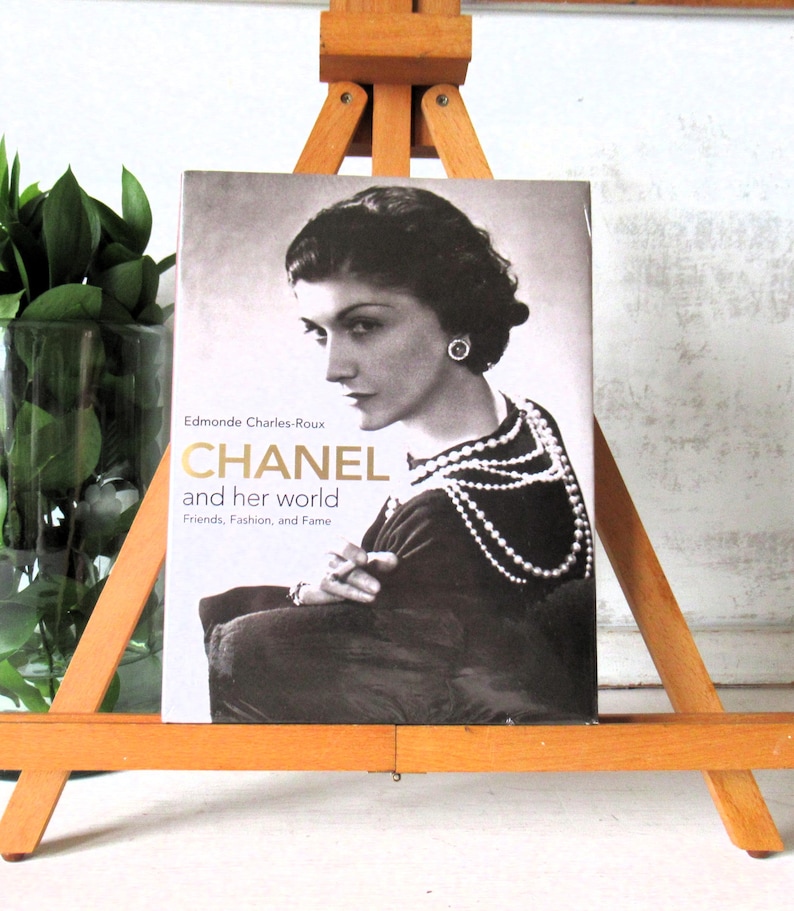 May include: A book titled "CHANEL and her world" by Edmonde Charles-Roux, featuring a black and white portrait of Coco Chanel. The book is displayed on a wooden easel, with a glass vase of green plants to the left.