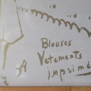 May include: A framed artwork featuring a sketch in gold tones on a light background. The text "Blouses, Vetements, imprim&eacute;s" is visible. The artwork has a simple, elegant style.