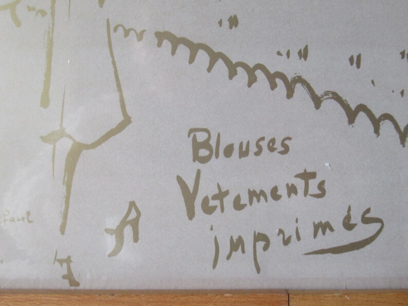 May include: A framed artwork featuring a sketch in gold tones on a light background. The text "Blouses, Vetements, imprim&eacute;s" is visible. The artwork has a simple, elegant style.