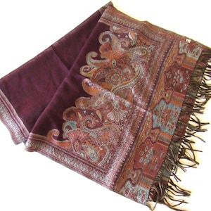 Puede incluir: Una bufanda de color ciruela con un borde de paisley detallado en tonos óxido, verde azulado y dorado. La bufanda tiene un borde con flecos y un cuerpo liso de color ciruela. Este accesorio es adecuado para añadir un toque de elegancia a cualquier atuendo.