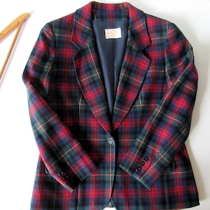 May include: A long-sleeve blazer featuring a red, navy blue, and dark green plaid pattern. The jacket has a notched lapel, a single front button closure, and three small buttons on each sleeve cuff. The interior is lined with solid navy blue fabric.