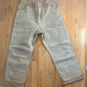 May include: Tan work pants featuring a double-front knee design with reinforced stitching, metal rivets at the pocket corners, a button closure at the waist, and a straight-leg cut with visible signs of wear, including small spots and minor fraying.
