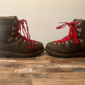 May include: A pair of dark brown leather boots featuring bright red laces, silver-toned metal eyelets and hooks, a padded collar, and a dark rubber sole. The side of the boots displays a circular embossed logo with the text Lowa.