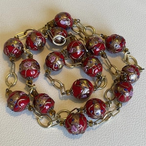 May include: A necklace featuring round red glass beads with gold-toned metallic flecks and small painted floral accents. The beads are connected by gold-toned metal chain links, creating a continuous circular design with a metallic clasp closure.