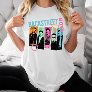 May include: A white short-sleeve t-shirt featuring a graphic print of the Backstreet Boys. The design includes five individual portraits of the band members set against colorful rectangular backgrounds, accented by neon geometric shapes and text.