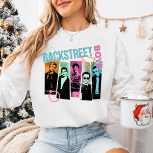 Peut inclure: Un sweat-shirt blanc à manches longues avec une impression graphique des Backstreet Boys. Le motif comprend cinq portraits individuels des membres du groupe sur des fonds rectangulaires colorés, avec le texte Backstreet Boys au-dessus.