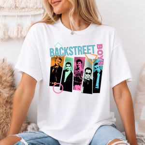 Pode incluir: Uma t-shirt branca de manga curta com uma impressão gráfica dos Backstreet Boys. O design inclui cinco retratos individuais dos membros da banda em painéis retangulares coloridos, acentuados por formas geométricas e o texto Backstreet Boys.