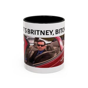 May include: A white ceramic mug with a black interior and rim, featuring a printed image of a person in a suit and sunglasses sitting in a red convertible car, with the black text IT'S BRITNEY, BITCH printed above the image on the mug surface.