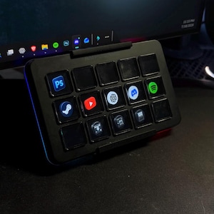 May include: A black macro keypad featuring fifteen square keys arranged in a grid. Several keys are illuminated with icons for software applications including Photoshop, Steam, YouTube, ChatGPT, Discord, and Spotify, set against a dark background.