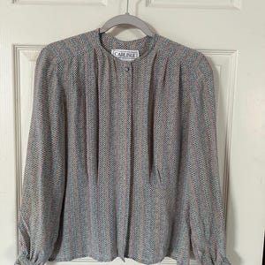 May include: A long-sleeved blouse with a herringbone pattern in gray, blue, and pink. The blouse has a round collar, a single button closure, and the brand name "CARLISLE" on a label.
