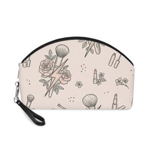 Puede incluir: Un neceser semicircular de color rosa pálido con un patrón repetitivo de ilustraciones de línea negra que muestran brochas de maquillaje, pintalabios, rosas y rayos, acabado con una cremallera negra y una correa de muñeca desmontable.