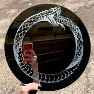 Könnte beinhalten: Ein runder schwarzer Spiegel mit einer weißen Gravur eines Ouroboros, einer Schlange, die ihren eigenen Schwanz frisst. Die detaillierte Darstellung zeigt die Schuppen der Schlange und das geöffnete Maul mit sichtbaren Zähnen in Ringform.