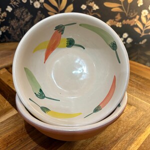 May include: A cream-colored ceramic bowl featuring a hand-painted pattern of chili peppers in shades of red, orange, yellow, and green. The interior of the bowl displays these colorful peppers scattered around the basin, while the exterior is a solid terracotta color.