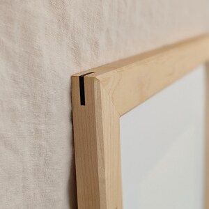 以下が含まれることがあります： 薄い色の木製フォトフレーム。留め継ぎのコーナー部分には、細く暗い垂直のインレイディテールが施されています。フレームは滑らかな天然木の木目仕上げで、中央の無地の白い長方形の領域を囲んでいます。