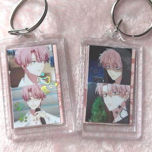 May include: Two rectangular clear acrylic keychains featuring anime-style illustrations of a character with pink hair. The left keychain displays two panels of the character, while the right keychain shows two different panels with holographic paw print accents.