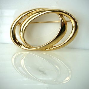 May include: A polished gold-tone metal brooch featuring two interlocking oval rings. The design consists of a smooth, reflective surface with a pin mechanism visible on the back. The brooch has a continuous, rounded shape with no additional embellishments.