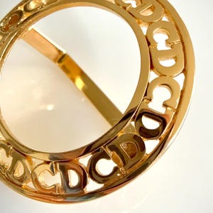 May include: A gold-toned circular ring featuring a repeating openwork pattern of the letters CD around the entire circumference. The metal has a polished, reflective finish, and the design consists of a continuous band of interconnected letters.
