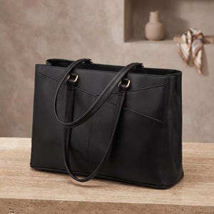 May include: A black structured tote bag with two long shoulder straps, gold-toned metal hardware, and a clean, minimalist exterior design featuring stitched panel detailing on the front. The bag has a rectangular silhouette and a flat base.