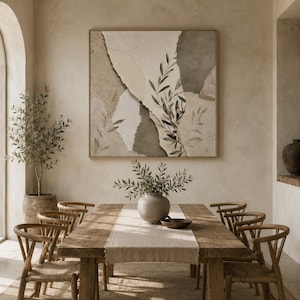 May include: A square wall art piece featuring abstract layered shapes in shades of beige, cream, and dark grey, accented with a painted olive branch motif. The artwork is framed in a thin light wood border and displayed on a neutral wall.