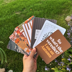 May include: A fan of rectangular cards with rounded corners. The top card is brown with white text reading So what's the verdict? I am an Autumn. Other cards feature black and white line art of a skull, brown stripes, and orange autumn-themed graphics.