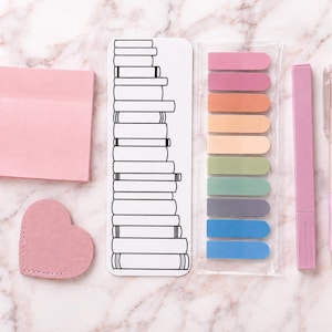 Puede incluir: Un conjunto de papelería que incluye un bloc de notas adhesivas cuadrado de color rosa claro, un marcapáginas de fieltro rosa en forma de corazón, un marcapáginas largo con una ilustración en blanco y negro de una pila de libros, nueve banderitas adhesivas de colores, un resaltador rosa rectangular y un bolígrafo retráctil rosa y blanco.