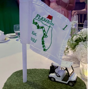 May include: A white golf flag with green text and a Florida outline, reading "Flemion Est. 2025" on a green turf base. A miniature white golf cart sits nearby. The flag is on a white pole.