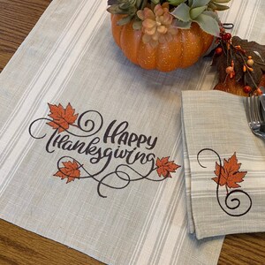 May include: A gray and white striped table runner with the text "Happy Thanksgiving" embroidered in black with orange leaves. A matching gray and white striped napkin with an orange leaf embroidered on it is on top of the table runner.