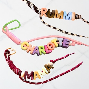 Op de afbeelding: Drie gepersonaliseerde koorden met kleurrijke letterkralen die RUMMI, CHARLOTTE en MAMA spellen. Elk koord is gemaakt van een patroon of effen koord, met een groene karabijnhaak en een gouden hartvormige sluiting.