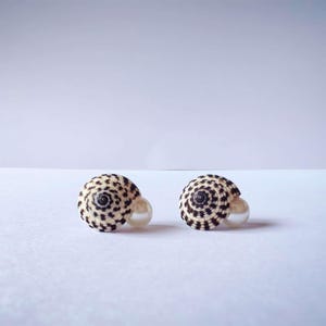 Puede incluir: Un par de pendientes de botón con conchas marinas en espiral con un patrón de manchas negras y blancas. Cada concha está acentuada con una cuenta de perla redonda de color crema colocada en la base, creando un contraste de textura y color.