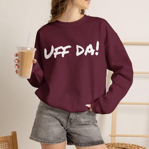 May include: A maroon long-sleeve crewneck sweatshirt featuring the white text UFF DA! printed across the chest in a bold, hand-lettered style font. The garment has a ribbed neckline, cuffs, and hem, providing a classic fit and solid color design.