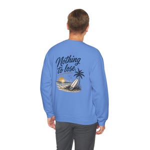 May include: A light blue crewneck sweatshirt featuring a graphic on the back. The design includes a palm tree, a surfboard leaning against it, a sunset over the ocean, and the text Nothing to lose. in a dark blue cursive script font.