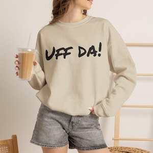May include: A beige long-sleeve crewneck sweatshirt featuring the text UFF DA! printed in black across the chest. The garment has a ribbed neckline, cuffs, and hem, and is paired with gray denim shorts. The design is simple and functional.