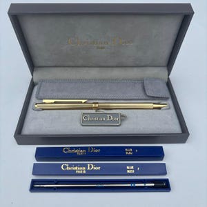May include: A gold-toned ballpoint pen with a ribbed barrel and a clip, presented in a grey hinged box with a grey velvet interior. The set includes a Christian Dior branded tag, two blue ink refill boxes labelled Christian Dior Paris, and one metal ink refill.