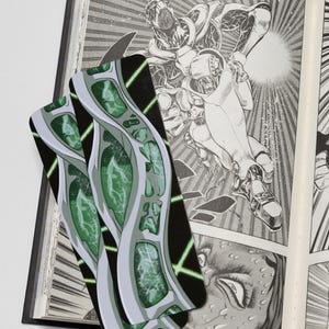 May include: Two rectangular bookmarks featuring a graphic design of interconnected silver metallic structures containing glowing green energy patterns, layered over an open book with black and white manga-style illustrations of a robotic figure.