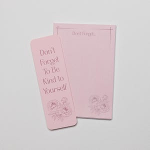 Puede incluir: Un marcapáginas rectangular de color rosa claro y un bloc de notas a juego. El marcapáginas presenta el texto "Don't Forget To Be Kind to Yourself" y una ilustración floral. El bloc de notas muestra el texto "Don't Forget..." y un diseño floral.