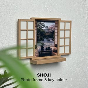 May include: A tan wall-mounted photo frame and key holder designed to resemble a shoji screen with two open panels. The central image depicts a Japanese garden scene with a table, tea set, and trees. The text SHOJI Photo frame & key holder is visible.