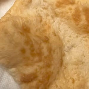 May include: A piece of fried flatbread with a golden-brown, bubbly, and uneven surface texture. The dough appears thin and cooked, showing variations in colour from light beige to deep tan across its surface, with visible air pockets.