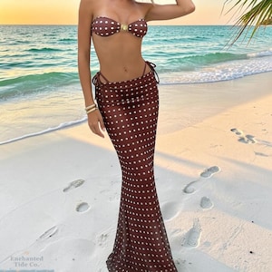 May include: A brown two-piece set featuring a strapless bandeau top with a gold-tone centre detail and a matching floor-length maxi skirt, both adorned with a white polka dot pattern. The skirt includes side tie closures at the waist.