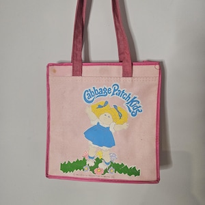 Pode incluir: Uma bolsa tote rosa com um gráfico Cabbage Patch Kids apresentando uma boneca loira com vestido azul, grama verde e flores coloridas. A bolsa tem alças e bordas rosa escuro, com o texto Cabbage Patch Kids e 1983 Original Appalachian Artworks, Inc.