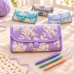 Könnte beinhalten: Ein rechteckiges Etui aus lavendelfarbenem Stoff mit Klappverschluss, gesichert durch einen passenden Knopf, verziert mit weißer Blumenstickerei. Drei Häkelnadeln mit violetten, grünen und pfirsichfarbenen Griffen liegen daneben, sowie weitere Etuis in Blau, Grau und Hellblau.