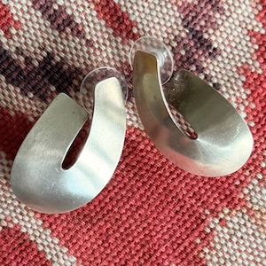 May include: A pair of silver-toned hoop earrings with a smooth, brushed metal finish and a wide, open U-shaped design. Each earring features a curved, flat surface that tapers slightly at the ends and includes a clear plastic backing for support.