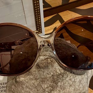 May include: Oversized sunglasses featuring a translucent amber-brown frame and dark brown gradient lenses. The frames have a rounded, wide shape with a clear bridge section connecting the two lenses, providing a classic look with warm tones.