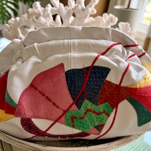 May include: A white leather clutch bag featuring a gathered top closure and a colorful geometric appliqué design. The surface is decorated with textured patches in shades of red, pink, dark blue, green, and yellow, accented by red stitching.