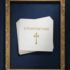 Può includere: Una pila di carte rettangolari bianche con angoli arrotondati, caratterizzate dal testo dorato 52 Scripture Cards e da un simbolo di croce dorata. Le carte sono posizionate all'interno di una cornice rettangolare dorata con dettagli decorati su una superficie blu scuro testurizzata.