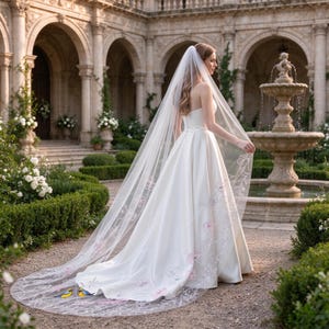 May include: A floor-length white wedding dress with a strapless bodice and a full skirt, paired with a long, sheer veil featuring delicate embroidered floral patterns in pink and green, along with small blue bird motifs near the hem.
