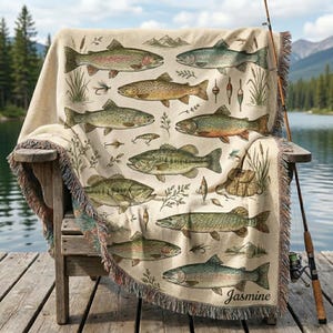 May include: A cream-colored woven throw blanket featuring a repeating pattern of various freshwater fish, including trout and bass, interspersed with fishing lures, reeds, and a satchel. The name Jasmine is embroidered in the bottom right corner.