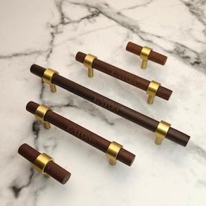 May include: A collection of five cabinet pulls and knobs made of dark brown wood with brushed gold metal accents. Each piece features the word EATON engraved on the wooden surface. The set includes two small cylindrical knobs and three long handles.
