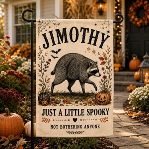 Könnte beinhalten: Eine rechteckige Gartenflagge mit einer zentralen Illustration eines Waschbären, der zwischen zwei Kürbissen läuft. Der Text lautet JIMOTHY, JUST A LITTLE SPOOKY und NOT BOTHERING ANYONE. Das Design enthält Spinnennetze, Fledermäuse und Herbstblätter.