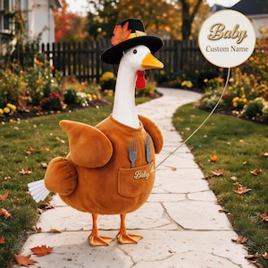 May include: A white goose figurine wearing a black pilgrim hat with an orange leaf, a brown fleece-like body covering, and orange feet. A pocket on the front holds a fork and knife, with the word Baby embroidered on it. A circular sign reads Baby Custom Name.